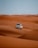 A white SUV is driving over vast orange sand dunes under a clear blue sky. The tracks of the vehicle are visible on the sand, suggesting movement and adventure.