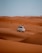 A white SUV is driving over vast orange sand dunes under a clear blue sky. The tracks of the vehicle are visible on the sand, suggesting movement and adventure.