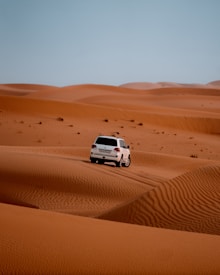 A white SUV is driving over vast orange sand dunes under a clear blue sky. The tracks of the vehicle are visible on the sand, suggesting movement and adventure.