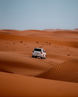 A white SUV is driving over vast orange sand dunes under a clear blue sky. The tracks of the vehicle are visible on the sand, suggesting movement and adventure.