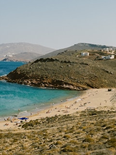 A scenic beach with turquoise water stretches alongside a sandy shore. People are relaxing and sunbathing on the beach, while others are swimming in the sea. The area is surrounded by rugged hills with sparse vegetation and a few small, white buildings atop them. The overall atmosphere suggests a serene and remote coastal location.