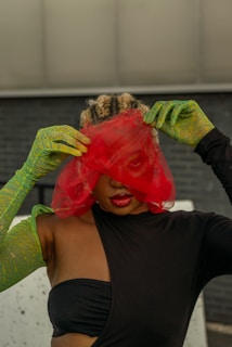 A person with braided hair is partially obscured by a red mesh veil. They are wearing a black outfit with one shoulder exposed, and long green gloves with a pattern. The background is a textured dark wall, adding contrast to the vibrant colors.