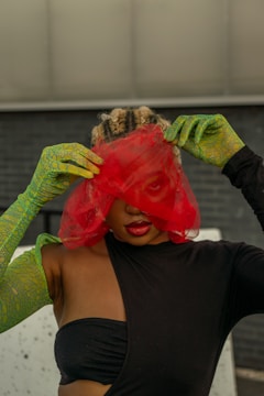 A person with braided hair is partially obscured by a red mesh veil. They are wearing a black outfit with one shoulder exposed, and long green gloves with a pattern. The background is a textured dark wall, adding contrast to the vibrant colors.