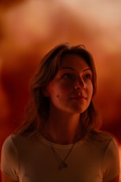 A person with long hair is gazing upwards with a contemplative expression. The background is a warm, blurred blend of orange and red tones, creating a serene and intimate atmosphere.