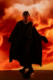 A person dressed in a dark, flowing outfit stands against a vibrant, fiery background that resembles swirling clouds or flames. The outfit includes platform shoes and a headpiece, adding a dramatic flair.