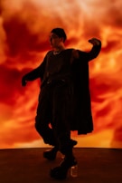 A person dressed in dark, edgy clothing strikes a dynamic pose against a vibrant, fiery background. They wear a beanie, sunglasses, sleeveless shirt, and cape-like garment, with their hand raised in a dramatic gesture. The overall scene is intense and expressive.