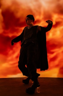 A person dressed in dark, edgy clothing strikes a dynamic pose against a vibrant, fiery background. They wear a beanie, sunglasses, sleeveless shirt, and cape-like garment, with their hand raised in a dramatic gesture. The overall scene is intense and expressive.