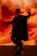 A person dressed in dark, edgy clothing strikes a dynamic pose against a vibrant, fiery background. They wear a beanie, sunglasses, sleeveless shirt, and cape-like garment, with their hand raised in a dramatic gesture. The overall scene is intense and expressive.