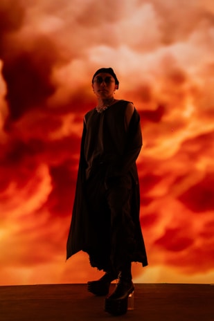 A person wearing stylish, dark clothing stands confidently against a dramatic, fiery orange background. The background resembles swirling clouds, creating a visually striking contrast with the person's attire. The setting appears theatrical, with a mood of mystery and intensity.