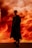 A person wearing stylish, dark clothing stands confidently against a dramatic, fiery orange background. The background resembles swirling clouds, creating a visually striking contrast with the person's attire. The setting appears theatrical, with a mood of mystery and intensity.