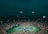 A large, illuminated tennis stadium filled with spectators during a nighttime match. The court is brightly lit, contrasting with the surrounding dark sky. Players are visible on the court, with a scoreboard in view showing match details.