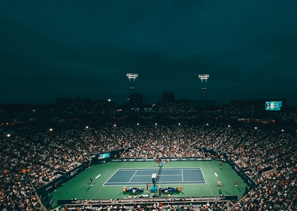 A large, illuminated tennis stadium filled with spectators during a nighttime match. The court is brightly lit, contrasting with the surrounding dark sky. Players are visible on the court, with a scoreboard in view showing match details.