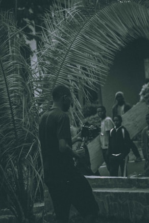 A group of people stands beneath large, arching palm fronds. One individual in the foreground is holding a camera on a stabilizer, while others in the background appear relaxed. The environment seems tropical, with lush greenery surrounding them.
