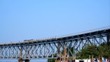 A large steel bridge spans horizontally across the image under a clear blue sky. The structure is supported by a complex network of beams and vertical pillars. Below the bridge, there are green trees and some poles, indicating a natural or open area. The bridge has some vehicles on it, which suggests it is in use.