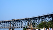 A large steel bridge spans horizontally across the image under a clear blue sky. The structure is supported by a complex network of beams and vertical pillars. Below the bridge, there are green trees and some poles, indicating a natural or open area. The bridge has some vehicles on it, which suggests it is in use.