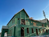 A building under construction is covered with green sheathing material. The roof is partially completed, and wooden framing is visible. The sky is clear and blue, and a tall communication tower is seen in the background.