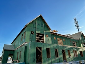 A building under construction is covered with green sheathing material. The roof is partially completed, and wooden framing is visible. The sky is clear and blue, and a tall communication tower is seen in the background.