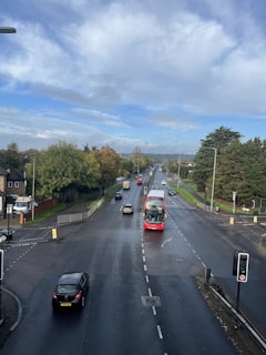 A clear road with a red double-decker bus driving through the center lane. Several other vehicles can be seen traveling in both directions. Lush green trees line the sides of the road, and there are traffic lights at the intersection. The sky is partly cloudy with patches of blue, and the road appears slightly wet, possibly after a recent rain.