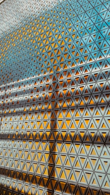 Geometric patterns formed by a series of triangular motifs create a grid-like structure. The interplay of light and shadow adds depth and complexity to the repetitive design, with vibrant shades of yellow and cool tones of blue dominating the color palette.