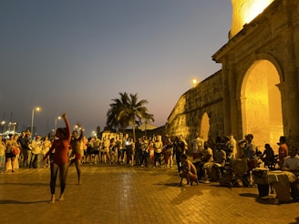 A group of people watching a street performance near an old stone structure illuminated by warm lights. A man and woman are dancing in the center, with live musicians playing drums on the right. Palm trees and a crowd of observers add to the lively atmosphere during dusk.