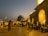 A group of people watching a street performance near an old stone structure illuminated by warm lights. A man and woman are dancing in the center, with live musicians playing drums on the right. Palm trees and a crowd of observers add to the lively atmosphere during dusk.