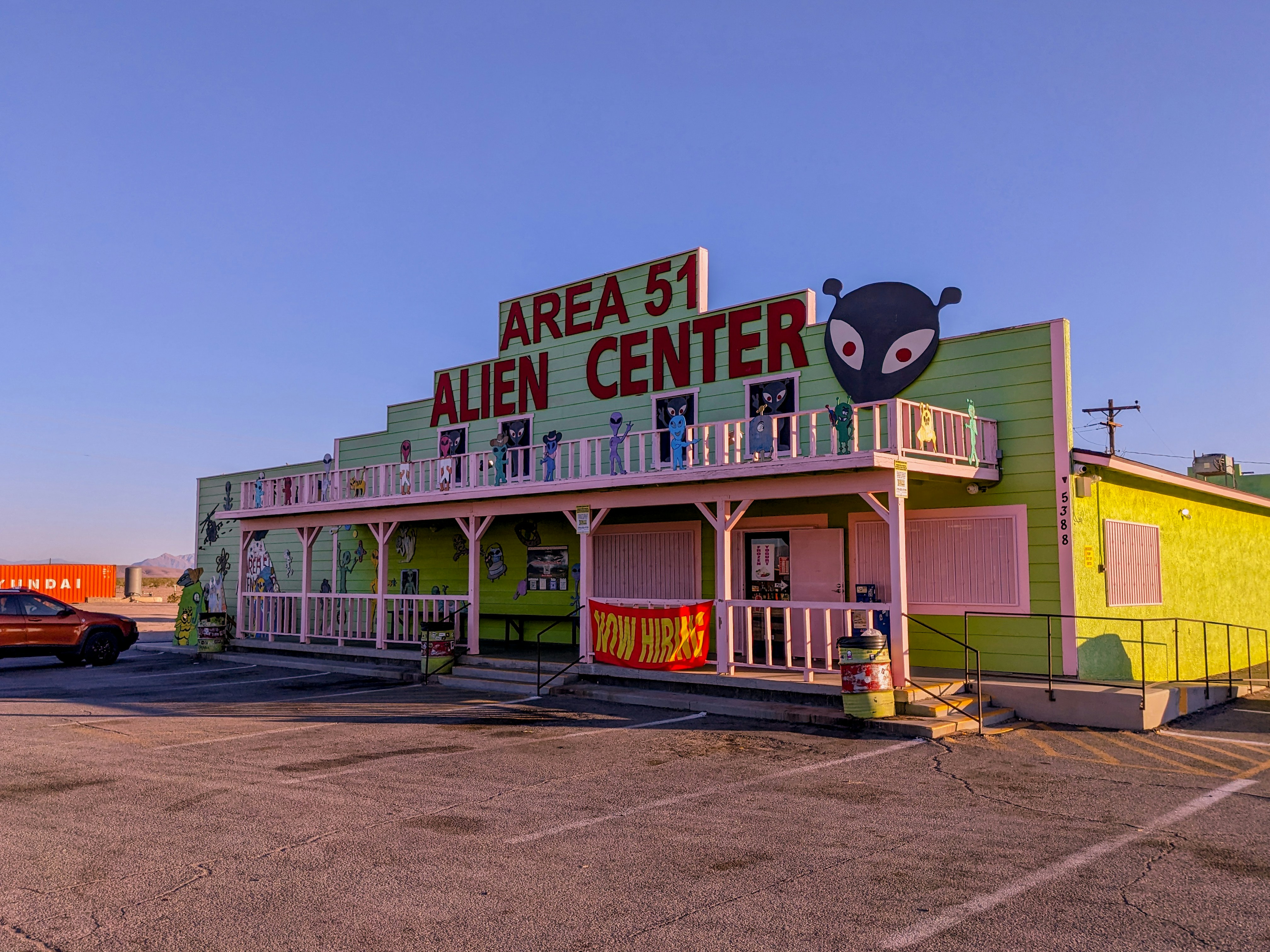 A building with a bright green exterior featuring a large sign that reads 'Area 51 Alien Center' in red. There is an alien head graphic on the right side of the sign. The building includes white trim and a balcony decorated with alien figures. Parked in front is an orange vehicle and to the side is a shipping container with the word 'Hyundai' visible.