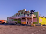 A building with a bright green exterior featuring a large sign that reads 'Area 51 Alien Center' in red. There is an alien head graphic on the right side of the sign. The building includes white trim and a balcony decorated with alien figures. Parked in front is an orange vehicle and to the side is a shipping container with the word 'Hyundai' visible.
