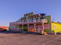 A building with a bright green exterior featuring a large sign that reads 'Area 51 Alien Center' in red. There is an alien head graphic on the right side of the sign. The building includes white trim and a balcony decorated with alien figures. Parked in front is an orange vehicle and to the side is a shipping container with the word 'Hyundai' visible.