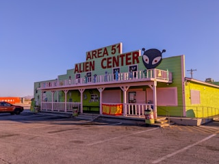 A building with a bright green exterior featuring a large sign that reads 'Area 51 Alien Center' in red. There is an alien head graphic on the right side of the sign. The building includes white trim and a balcony decorated with alien figures. Parked in front is an orange vehicle and to the side is a shipping container with the word 'Hyundai' visible.