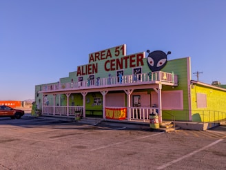 A building with a bright green exterior featuring a large sign that reads 'Area 51 Alien Center' in red. There is an alien head graphic on the right side of the sign. The building includes white trim and a balcony decorated with alien figures. Parked in front is an orange vehicle and to the side is a shipping container with the word 'Hyundai' visible.