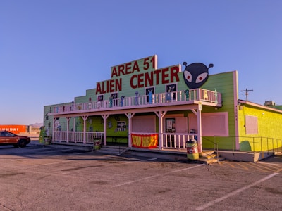 A building with a bright green exterior featuring a large sign that reads 'Area 51 Alien Center' in red. There is an alien head graphic on the right side of the sign. The building includes white trim and a balcony decorated with alien figures. Parked in front is an orange vehicle and to the side is a shipping container with the word 'Hyundai' visible.
