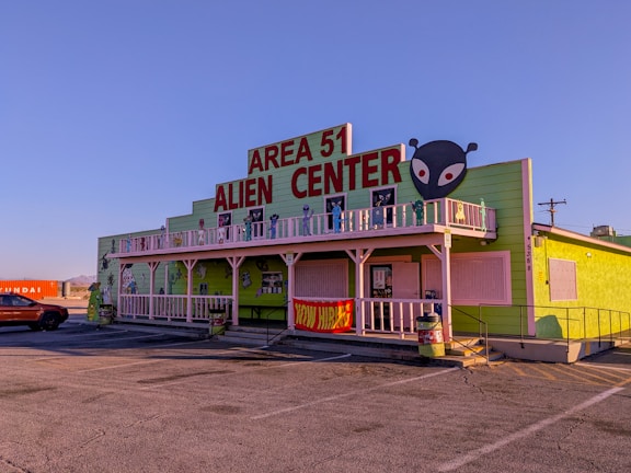 A building with a bright green exterior featuring a large sign that reads 'Area 51 Alien Center' in red. There is an alien head graphic on the right side of the sign. The building includes white trim and a balcony decorated with alien figures. Parked in front is an orange vehicle and to the side is a shipping container with the word 'Hyundai' visible.