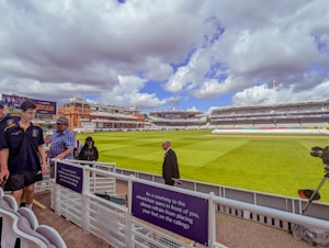 A cricket stadium with a well-maintained green field under a partly cloudy sky. Several people are visible near the stands, wearing casual and sports attire. The red-brick architecture of the stadium's building is prominent in the background.