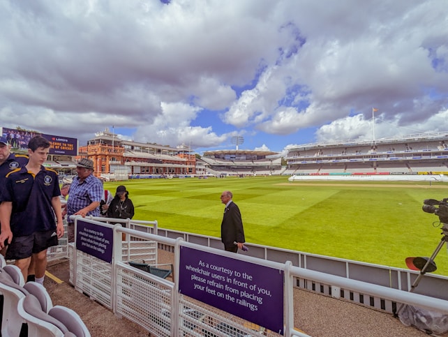 A cricket stadium with a well-maintained green field under a partly cloudy sky. Several people are visible near the stands, wearing casual and sports attire. The red-brick architecture of the stadium's building is prominent in the background.