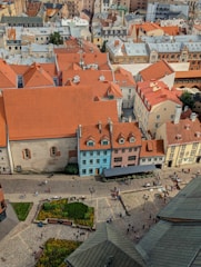 An aerial view of a European cityscape featuring a collection of buildings with orange-tiled roofs. The structures vary in heights and styles, showcasing both modern and traditional architecture. There is a central square with cobblestone streets, flower beds, and people walking and socializing. A few benches and street lamps are scattered around the square.