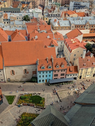 An aerial view of a European cityscape featuring a collection of buildings with orange-tiled roofs. The structures vary in heights and styles, showcasing both modern and traditional architecture. There is a central square with cobblestone streets, flower beds, and people walking and socializing. A few benches and street lamps are scattered around the square.