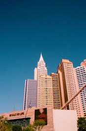 A clear blue sky provides the backdrop for a cluster of tall, modern skyscrapers with varied architectural designs. In the foreground, a building facade features a large advertisement that includes the phrase 'NO SALT NO LIFE' and an image of food. The bright sunlight casts a warm glow on the buildings.