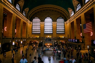 A grand hall with high ceilings, large arched windows, and ornate architectural details. The floor is bustling with people walking or standing, and there is an information booth in the center. An American flag is prominently displayed on the right side, and there is warm artificial lighting that highlights the stone walls.