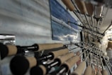 A close-up shot of several fishing rods displayed in a row on a wall, with their handles in focus and their tips pointing towards the ceiling. A picture featuring someone fishing at sea is mounted on the wall among the rods. The background shows part of an indoor space with a person visible in the distance.