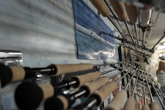 A close-up shot of several fishing rods displayed in a row on a wall, with their handles in focus and their tips pointing towards the ceiling. A picture featuring someone fishing at sea is mounted on the wall among the rods. The background shows part of an indoor space with a person visible in the distance.