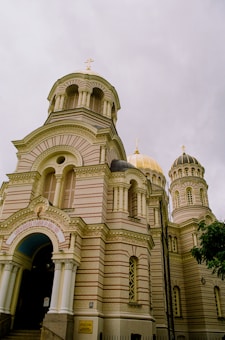 A grand Orthodox church with stunning architecture, featuring multiple domes and intricate detailing on its fa&ccedil;ade. The building is adorned with decorative patterns and ornate columns, and its exterior predominantly consists of beige and light brown bricks. The central dome is topped with a golden cross, adding to the regal appearance.
