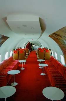 The interior of a vintage airplane features a long aisle lined with plush red seating on both sides. Each seat section includes white round tables in front of the seating areas. The design is reminiscent of mid-20th century luxury, with a spacious cabin and minimalist decor.