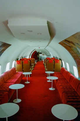 The interior of a vintage airplane features a long aisle lined with plush red seating on both sides. Each seat section includes white round tables in front of the seating areas. The design is reminiscent of mid-20th century luxury, with a spacious cabin and minimalist decor.