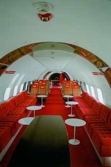 The interior of a retro-styled airplane cabin, featuring red plush seating along both sides and several small white circular tables. The seats have a vintage plaid pattern, and the decor appears luxurious and mid-century modern. Windows line both walls, allowing light to illuminate the space minimally.