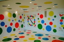 The image features a room decorated with colorful circles on the walls, floor, and ceiling in different sizes and colors. There is a large Twister game spinner on one wall and instructions for the game displayed on two walls. The circles are primarily red, yellow, green, and blue. The room has a playful and fun atmosphere.