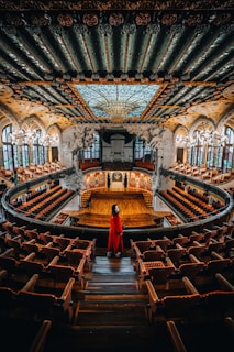 A grand concert hall features an intricately designed ceiling with ornate patterns and a large stained glass dome. The hall is adorned with rows of red cushioned seats and a wooden stage at the front. A person in a striking red outfit stands in the aisle, facing the stage.