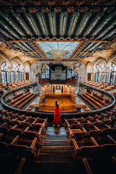 A grand concert hall features an intricately designed ceiling with ornate patterns and a large stained glass dome. The hall is adorned with rows of red cushioned seats and a wooden stage at the front. A person in a striking red outfit stands in the aisle, facing the stage.