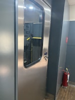 A metallic door with a glass window is seen in a corridor with gray walls and a wooden floor. Next to the door, there is a red fire extinguisher.