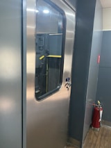 A metallic door with a glass window is seen in a corridor with gray walls and a wooden floor. Next to the door, there is a red fire extinguisher.