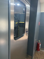 A metallic door with a glass window is seen in a corridor with gray walls and a wooden floor. Next to the door, there is a red fire extinguisher.